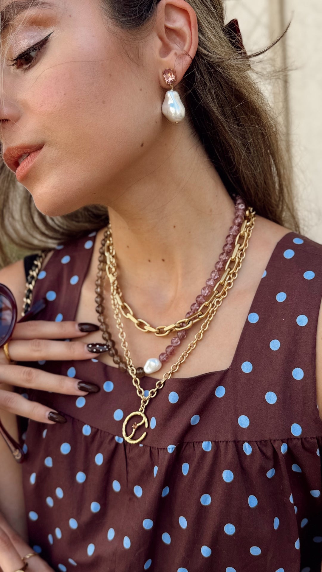 A close-up lifestyle shot of a woman wearing a layered gold necklace with a double-O pendant, a pink beaded strand, and a white pearl, paired with a brown and blue polka-dot top.