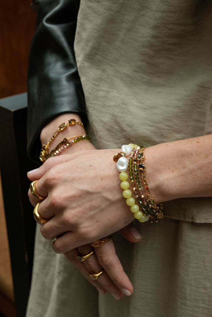 A close-up of a person's hands and wrists wearing multiple layered bracelets. The bracelets include gold-tone chains, beaded strands in yellow, green, and brown, and a white pearl.