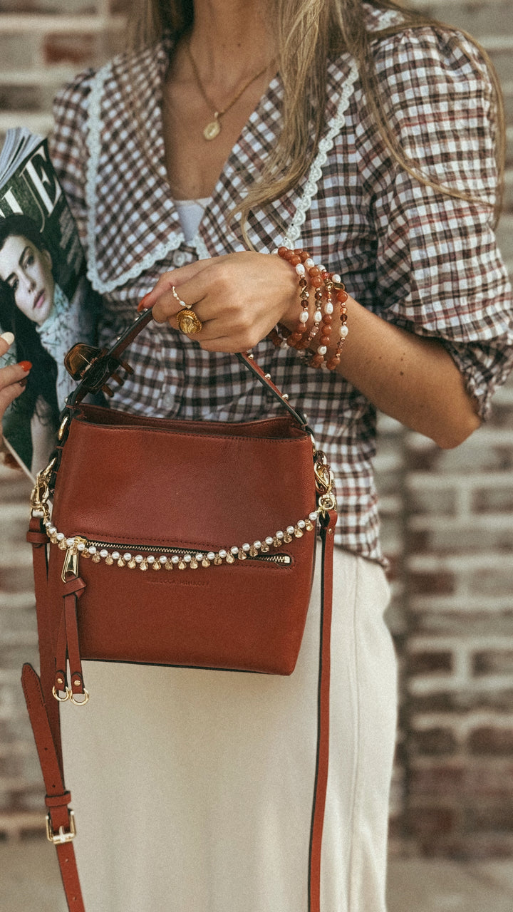 Person wearing a plaid shirt and holding a brown handbag against a brick wall.