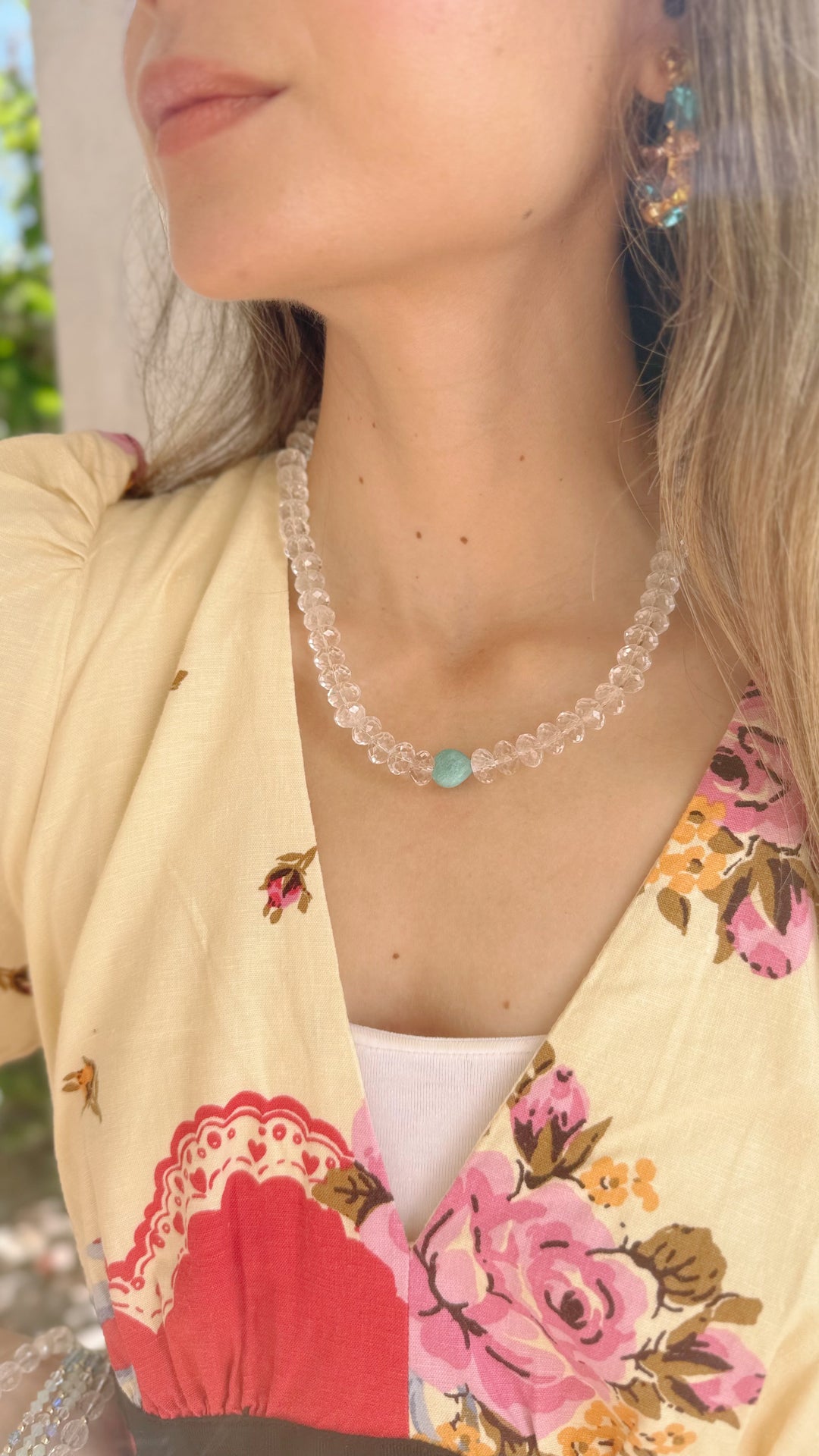 Person wearing a floral top and a clear quartz necklace with a blue gemstone.