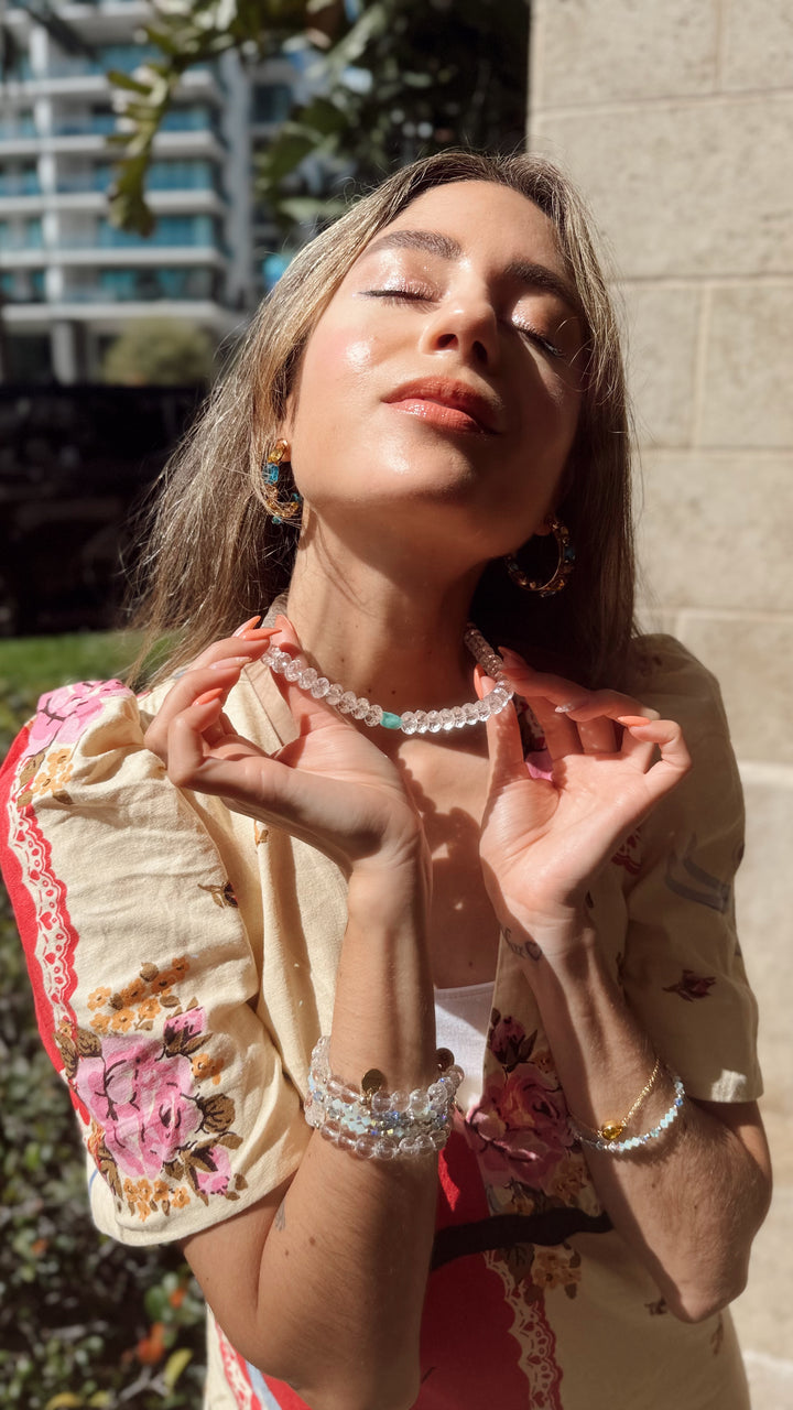 Woman wearing a floral dress and jewelry outdoors with sunlight casting shadows.